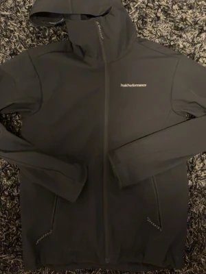Svart vindjacka från Peak Performance - Svart vindjacka med huva från Peak Performance. Jackan har hel dragkedja framtill, två fickor med dragkedja och diskret logga på bröstet. Tillverkad i ett lätt och smidigt material som passar perfekt för blåsiga dagar.