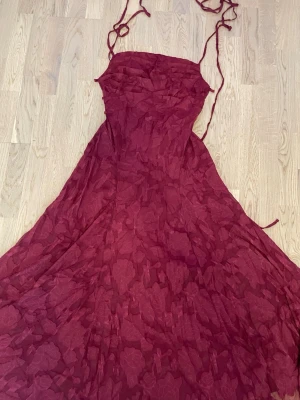 Vinröd långklänning med blommönster - Elegant vinröd långklänning med tunna axelband och subtilt blommönster. Klänningen har en figurnära överdel och ett svepande fall som ger en romantisk känsla. Perfekt för speciella tillfällen där du vill sticka ut.