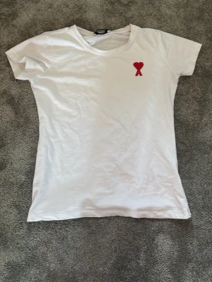 Vit t-shirt med rött hjärta från ami Paris  - Vit t-shirt från ami paris med ett litet rött hjärta och bokstaven A broderat på bröstet. Klassisk passform och rund halsringning. Perfekt basplagg med en enkel men snygg detalj framtill.