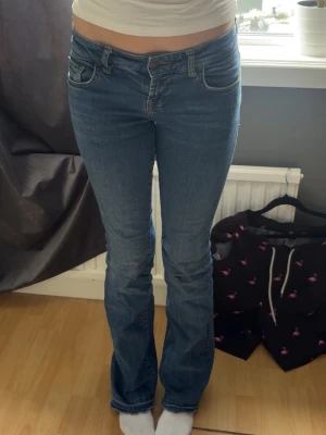 Low waist Blå bootcut jeans  - Säljer ett par klassiska blå bootcut jeans med fem fickor. Jeansen har en snygg tvätt och kontrastsömmar, samt broderade detaljer på bakfickorna. Perfekta för en avslappnad och trendig look. Modellen är 170cm lång. storlek 26/34. Har en liten fläck som säkert går bort i tvätt!