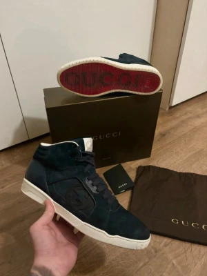 Gucci Rebound - Tja, Säljer ett par mörkblå Gucci skor mocka med vit sula och tydlig logga på sidan och på den röda sulan. Storlek 44, men passar nog också 45. Skorna har snörning, diskreta vita detaljer och röd undersida med Gucci-tryck. Nästan helt nya med inga slitningar någonstans. Dom är mid top, alltså inte så höga ändå. Box och dustbag medföljer tyvärr ej, men skickas i en ersättnings box.                                                                             Hör av er vid frågor eller funderingar! 