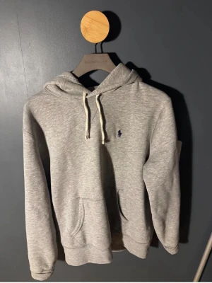 Grå hoodie från Polo Ralph Lauren - Snygg gråmelerad hoodie från Polo Ralph Lauren med klassisk huva och vita dragsnören. Har en känguruficka framtill och den ikoniska lilla blå loggan broderad på bröstet. Tillverkad i mjukt bomullsmaterial som är perfekt för chill dagar.