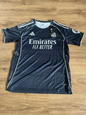 Real Madrid bortatröja Adidas mörkblå - Snygg mörkblå Real Madrid bortatröja från Adidas med vita detaljer och klubbmärke på bröstet. Tröjan har korta ärmar, rund hals och är tillverkad i ett lätt, ventilerande funktionsmaterial. Perfekt för fotbollsträning eller att bära till vardags.