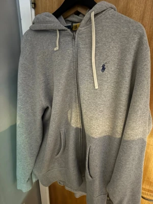 Grå hoodie från Polo Ralph Lauren - Snygg grå zip-hoodie från Polo Ralph Lauren med klassisk blå logga på bröstet. Hoodien har dragsko i huvan, två fickor framtill och är tillverkad i mjukt bomullsmaterial. Perfekt för en chill och avslappnad stil.