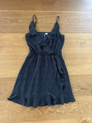 Svart prickig klänning från H&M Divided - Söt svart kortklänning från H&M Divided med vita prickar, smala axelband och volangkant nedtill. Klänningen har omlott-look upptill och markerad midja för en feminin siluett. Perfekt för varma dagar och somriga tillfällen.