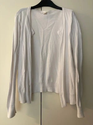 Vit basic kofta från H&M - En enkel och stilren vit kofta från H&M Basic. Koftan har knappar framtill, har långa ärmar och ribbade muddar vid ärmslut och nederkant. Perfekt att slänga över en topp för en clean look. Tillverkad i mjukt material.