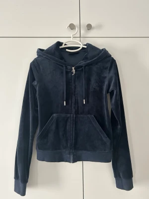 Mörkblå velour hoodie Juicy Couture - Mjuk och lyxig mörkblå hoodie från Juicy Couture i velourmaterial. Dragkedja framtill, två stora fickor och justerbar huva med snören. Perfekt för en avslappnad och trendig look.