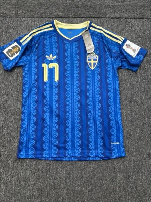Blå Sverige fotbollströja Adidas - Säljer en blå fotbollströja från Adidas med Sveriges landslagsemblem och nummer 17 Gyökeres. Tröjan har gula detaljer, mönstrad design och korta ärmar. Perfekt för dig som vill stötta Sverige på match eller träning.