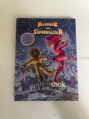 Handbok för superhjältar: Jullovsbok - Handbok för superhjältar: Jullovsbok är en färgglad och illustrerad bok med vintertema. Omslaget visar två superhjältar i röda och gula dräkter som svävar över ett snötäckt stadslanskap. Boken innehåller pyssel, äventyr och juliga utmaningar.