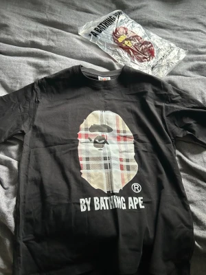 Svart BAPE t-shirt med rutig logga - Säljer en svart t-shirt från BAPE med den klassiska apansiluetten i rutigt mönster på bröstet och texten 'BY BATHING APE' under. T-shirten har rund hals och korta ärmar. Detaljerad etikett med BAPE-logga på ärmen. Perfekt för dig som gillar streetwear.
