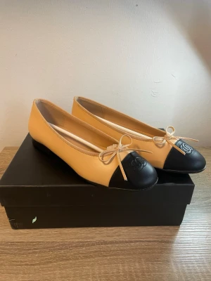 Beige och svarta ballerinas från Chanel - Snygga ballerinas från Chanel i beige skinn med svart tåparti och klassisk rosettdetalj framtill. Ikoniska CC-loggan är präglad på tån. Perfekta för dig som vill ha en tidlös och elegant look med lyxig känsla.