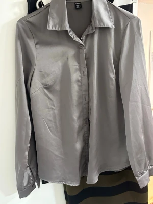 Silvrig skjorta i satinlook - Stilren silvrig skjorta från H&M med klassisk krage och knappar framtill. Skjortan har lång ärm och är tillverkad i ett glansigt satinliknande material som ger en lyxig känsla. Perfekt för dig som vill sticka ut med en elegant touch.