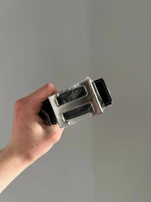 Svart skärp med stor silverspänne - Snyggt svart skärp med ett stort, ikoniskt H-format silverspänne. Skärpet har vita sömmar längs kanterna och är tillverkat i skinn. Perfekt accessoar för att lyfta vilken outfit som helst.