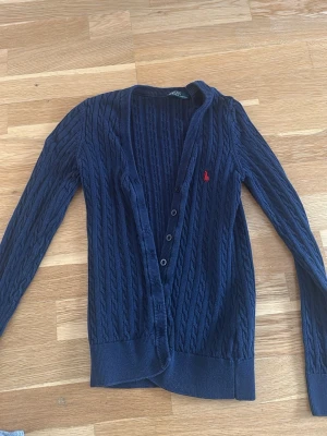 Mörkblå kabelstickad kofta Polo Ralph Lauren - Kabelstickad mörkblå kofta från Polo Ralph Lauren med knappar framtill och röd broderad logga på bröstet. Långärmad modell med ribbade muddar och klassisk v-ringad öppning. Perfekt för lager på lager och snygg till jeans.