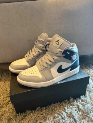 Air Jordan 1 Mid grå/vit/marin - Snygga Air Jordan 1 Mid sneakers i grått, vitt och marinblått läder. Klassisk siluett med perforerad tå, ikonisk Swoosh och Jumpman-logga på plösen. Skorna har högt skaft och vita snören. Perfekta för dig som gillar streetwear och basketstil.