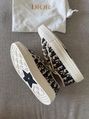 Dior Oblique låga sneakers Walk’n’dior 37 - Snygga låga sneakers från Dior med klassiskt blått och beige oblique-mönster. Skorna har vita gummisulor, beige snörning och tydlig 'Christian Dior'-logga på plösen och sidan. Ovandelen är i textil med exklusiv känsla och detaljer i vitt. Perfekta för en trendig streetwear-look.