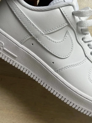 Nike Air Force 1 vita sneakers - Klassiska helvita Nike Air Force 1 sneakers i lågt utförande. Tillverkade i slittåligt läder med perforerad tåbox för ventilation, kraftig sula och ikoniska Swoosh-loggan på sidan. Skorna har vita skosnören och metallbricka med AF1-detalj.