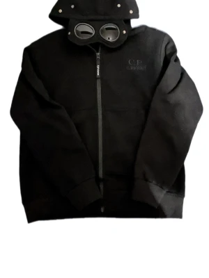 Svart hoodie med goggles från C.P. Company - Svart hoodie från C.P. Company med hel dragkedja och ikonisk huva med inbygga goggles. Tröjan är knappt använd och är i väldigt bra skick men säljer då den inte passar tyvärr