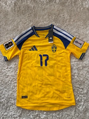 Sverige matchtröja Adidas gul/blå - Gul och blå matchtröja från Adidas med Sveriges landslagsemblem och nummer 17 på bröstet. Tröjan har blå detaljer, vita ränder på axlarna och officiella turneringsmärken på ärmarna. Tillverkad i lätt funktionsmaterial med Heat.RDY-teknologi.