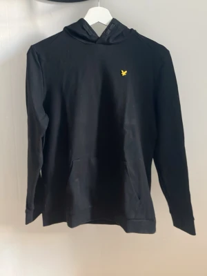Svart hoodie från Lyle & Scott - Snygg svart hoodie från Lyle & Scott med klassisk gul logga på bröstet. Hoodien har huva, känguruficka och långa ärmar. Tillverkad i mjukt bomullsmaterial som är skönt att ha på sig. Perfekt för en avslappnad och stilren look. Priset går såklart att diskutera 😁