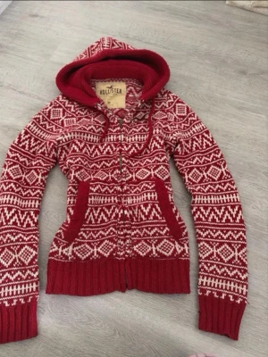 Rödvit stickad hoodie från Hollister - Säljer en röd och vit stickad hoodie från Hollister med dragkedja och snyggt mönster. Tröjan har huva med dragsko, två fickor fram och ribbade muddar. Perfekt för kalla dagar och har en broderad Hollister-logga på bröstet.