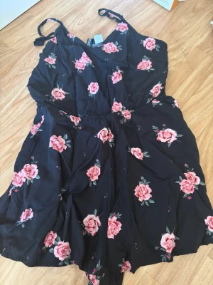 Svart blommig jumpsuit med rosor - Supersöt svart jumpsuit med rosa rosor och tunna axelband. Har en markerad midja med resår och korta ben. Perfekt för sommardagar och enkel att styla med sneakers eller sandaler. Materialet känns lätt och luftigt.