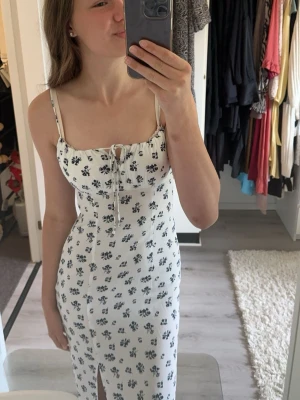 Vit blommig långklänning med snörning - Supersöt långklänning i vit med blått blommönster och smala axelband. Klänningen har snörning framtill vid bysten och en liten slits framtill. Perfekt för sommarens alla tillfällen och ger en fräsch och romantisk vibe.