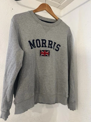 Grå sweatshirt från Morris - Snygg grå sweatshirt från Morris med mörkblå text och en broderad brittisk flagga på bröstet. Tröjan har rund halsringning, ribbade muddar och är tillverkad i mjukt bomullsmaterial. Perfekt för en avslappnad och stilren look.