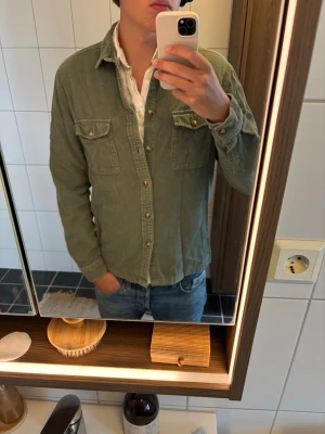 Grön manchester overshirt från ASOS - Säljer en grön overshirt i manchester från ASOS DESIGN. Skjortan har två bröstfickor med knappar, klassisk krage och knäppning framtill. Perfekt att bära öppen över en t-shirt eller stängd som skjorta. Snygg och avslappnad stil.