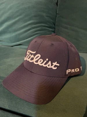 Mörkblå Titleist keps Pro V1 - Snygg mörkblå keps från Titleist med broderad logga framtill och texten Pro V1 på sidan. Justerbar rem baktill och patch med Titleist-logga. Klassisk böjd skärm och ventilerande hål. 