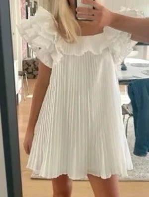 Zara klänning - Supersöt vit kortklänning med plisserad kjol och stora volangdetaljer vid axlarna. Klänningen har en luftig passform och är perfekt för varma dagar. Den fyrkantiga ringningen ger en trendig touch och materialet känns lätt och bekvämt.