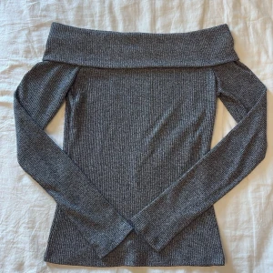 Grå off shoulder stickad tröja - Säljer en snygg grå ribbstickad off shoulder-tröja med långa ärmar. Tröjan har en bred krage som viks ner över axlarna och är figurnära i modellen. Använd fåtal gånger.