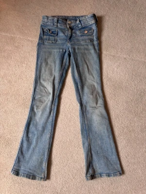 Ljusblå bootcut jeans med fickdetaljer - Säljer ett par ljusblå bootcut jeans med dubbla knappar i midjan och snygga fickor med lock och knapp framtill. Jeansen har en klassisk tvättad look och är tillverkade i mjukt denimtyg. Perfekta för en avslappnad och trendig stil.