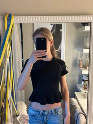 Svart croppad topp med volang - Trendig svart croppad topp med korta ärmar och volangkant nedtill. Toppen har en tight passform och är tillverkad i ett stretchigt material som ger en snygg siluett. Perfekt att styla med höga jeans eller kjol.