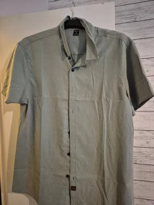 Ljusgrön kortärmad skjorta H&M - Stilren ljusgrön skjorta från H&M med korta ärmar och klassisk krage. Skjortan har knäppning framtill med svarta knappar och är tillverkad i ett mjukt, tunt tyg som passar perfekt till sommaren. Enkel och clean design utan fickor eller tryck.