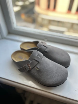 Grå Birkenstock Boston tofflor - Säljer ett par grå Birkenstock Boston tofflor i mocka med justerbart spänne i metall. Klassisk slip-in modell med mjuk fotbädd och korksula för extra komfort. Perfekta för dig som gillar stilrena och bekväma skor.