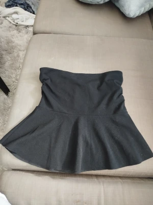 Svart bandeau tubtopp med volang - Stilren svart bandeau tubtopp med bred volang nedtill som ger en peplum-effekt. Toppen är axelbandslös och har en enkel, trendig look. Använd endast 1 gång 
