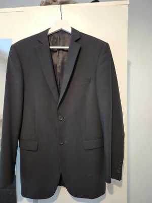 Svart kavaj från Dressmann - Stilren svart kavaj från Dressmann i modellen 42 Regular. Kavajen har två knappar framtill, klassiska slag och tre knappar vid ärmsluten. Tillverkad i ett stretchigt ullmaterial för extra komfort. Perfekt för en snygg och modern look.