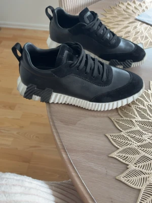H skor  - Säljer ett par svarta sneakers med chunky vit sula och coola detaljer på sidan. Skorna har en mix av mocka och slätt material, samt svarta skosnören. Perfekta för dig som gillar streetstyle och vill sticka ut lite extra.