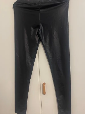 Svarta glansiga leggings S/M - Säljer ett par svarta leggings med glansig finish. De är tillverkade i 89% polyester och 11% elastan vilket gör dem stretchiga och bekväma. Perfekta för dig som gillar en trendig och edgy look. Midjehöga och med åtsittande passform.