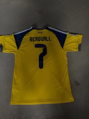 Svensk landslagströja Adidas nr 7 - Gul svensk landslagströja från Adidas med blå detaljer och nummer 7 på både fram- och baksida. Namnet 'Bergvall' står på ryggen. Tröjan har officiella landslagsmärken och patchar på ärmarna. Perfekt för fotbollsfans som vill stötta Sverige. Helt ny replika