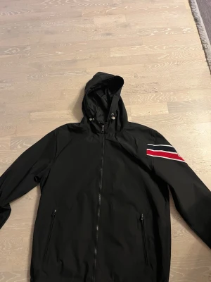 Svart Moncler vindjacka med huva - Snygg svart vindjacka från Moncler med huva och dragkedja framtill. Jackan har två fickor med dragkedja och en cool detalj med röd, vit och marinblå rand på ena ärmen samt Moncler-logga. Perfekt för dig som vill ha en stilren och sportig look.