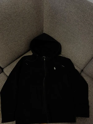 Ralph Lauren - Säljer en svart hoodie med dragkedja från Polo Ralph Lauren. Klassisk design med huva och två fickor framtill. Liten vit logga broderad på bröstet. Tillverkad i mjukt bomullsmaterial som är skönt att ha på sig. Perfekt för en avslappnad och stilren look.