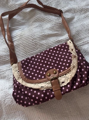 Brun axelväska med prickigt mönster - Söt brun axelväska med vita prickar och dekorativ spetskant. Väskan har ett lock med spänne i brunt konstläder och justerbar axelrem. Perfekt för dig som gillar vintage-inspirerad stil och vill ha en unik accessoar.