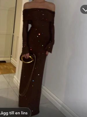 Meshki Brun glittrig offshoulder långklänning - Snygg brun långklänning från Meshki med glittriga detaljer och offshoulder-design. Klänningen har långa ärmar och en figurnära passform som framhäver siluetten. Perfekt för dig som vill sticka ut med en elegant och trendig look.