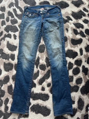 Mörkblåa bootcutjeans från True Religion - Säljer ett par mörkblåa bootcut jeans från True Religion med låg midja, i storlek 24. Lite långa för mig som är ca 162 cm. Säljer pga de tyvärr blivit för små för mig. Jag köpte jeansen här via plick men de har inte kommit till användning mycket i min ägo. Skicket är bra utöver slitningar längst ner på benen (se bild 3)😁
