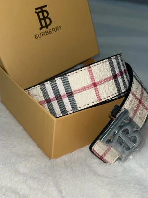 Burberry rutigt bälte med silverdetalj - Snyggt Burberry-bälte med klassiskt rutigt mönster i vitt, svart och rött. Bältet har ett stort silvrigt spänne med Burberrys logotyp. Perfekt accessoar för att lyfta vilken outfit som helst.