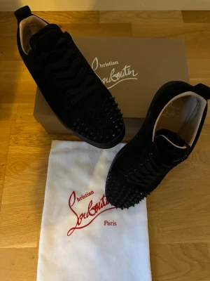 Svarta Christian Louboutin sneakers - Snygga svarta sneakers från Christian Louboutin med klassiska röda sulor och svarta nitar på tån. Skorna är låga och har en lyxig känsla med mockaliknande material och svarta skosnören. Perfekta för dig som vill sticka ut med exklusiva detaljer.