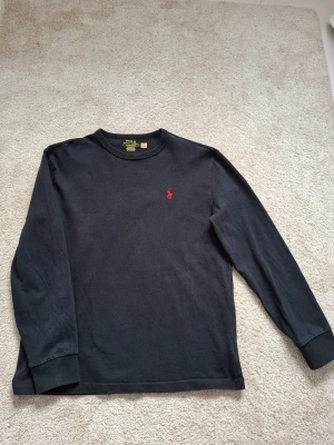 Svart långärmad tröja Polo Ralph Lauren - Klassisk svart långärmad tröja från Polo Ralph Lauren med rund halsringning och röd broderad logga på bröstet. Tröjan har ribbade muddar vid ärmslut och är tillverkad i mjuk bomull för en skön känsla.
