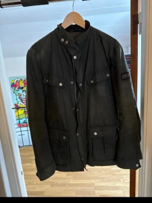Mörkgrön jacka från Barbour - Klassisk mörkgrön jacka från Barbour med fyra framfickor, hög krage och tryckknappar. Jackan har dragkedja och knappar framtill samt Barbour-logga på ärmen. Perfekt för dig som gillar tidlös och funktionell stil.